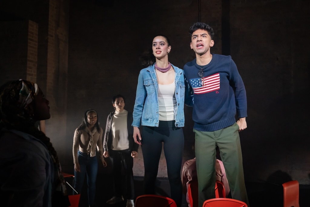 Action shot of the spring 2024 Shakespeare in Performance at RADA final presentation of Julius Caesar. Two actors in mid-performance. The actor on the right wears a sweater with an American flag stitched on the front and delivers a monologue while standing beside another actor. Some additional actors look on from behind the two featured actors.